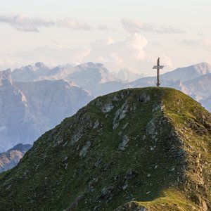 Gipfel mit großem Kreuz vor Berglandschaft im Sonnenuntergang