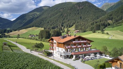 Alpenhotel mit Terrasse umgeben von grünen Wiesen und Bergen bei sonnigem Wetter
